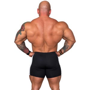 A muscular man with tattoos on his arms and legs stands with his hands on his hips, showcasing his well-defined physique.