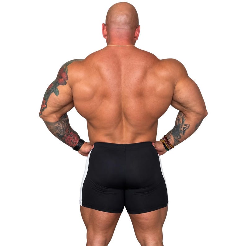 A muscular man with tattoos on his arms and legs stands with his hands on his hips, showcasing his well-defined physique.