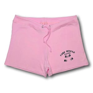 Puede incluir: Pantalones cortos deportivos de color rosa claro con cintura el&aacute;stica y cord&oacute;n a juego. La parte inferior de la pierna derecha muestra un estampado gr&aacute;fico negro de dos mancuernas con el texto CAMP MUSCLE BODYWEAR arqueado encima.