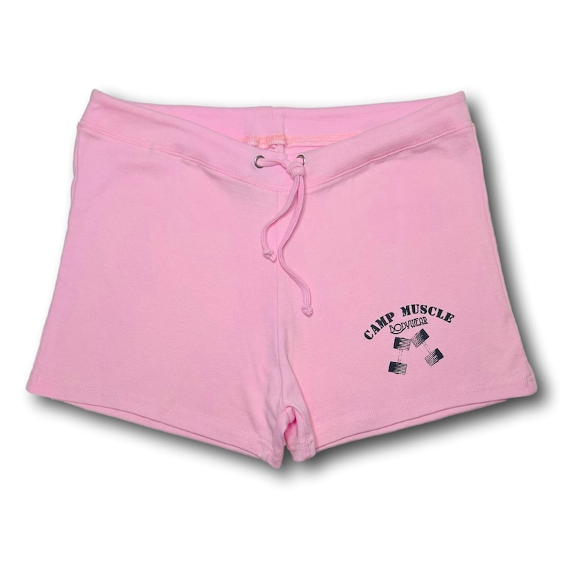 Puede incluir: Pantalones cortos deportivos de color rosa claro con cintura el&aacute;stica y cord&oacute;n a juego. La parte inferior de la pierna derecha muestra un estampado gr&aacute;fico negro de dos mancuernas con el texto CAMP MUSCLE BODYWEAR arqueado encima.