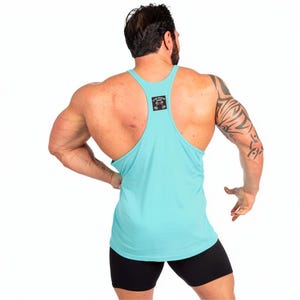 May include: A light turquoise racerback tank top featuring a small black square patch on the upper back with a white graphic of two dumbbells and the text "TEAM MUSCLE GYM" printed in white lettering, paired with black athletic shorts.