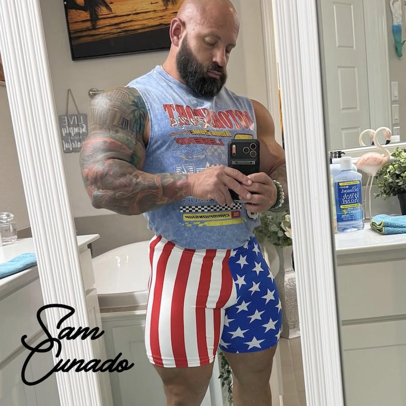May include: A person wearing a light blue tank top with a graphic design and red, white, and blue shorts with a star and stripe pattern. The person is holding a phone and looking in a mirror.