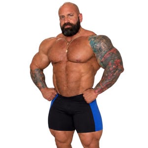 A muscular man with a beard and tattoos posing confidently in black shorts against a white background.