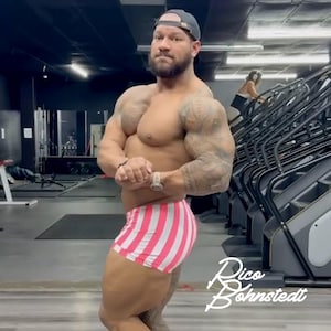 May include: A muscular person wearing pink and white striped shorts and a dark baseball cap poses in a gym. The person has tattoos on their arms and a watch on their wrist. The gym has exercise equipment and a person on a stair stepper.