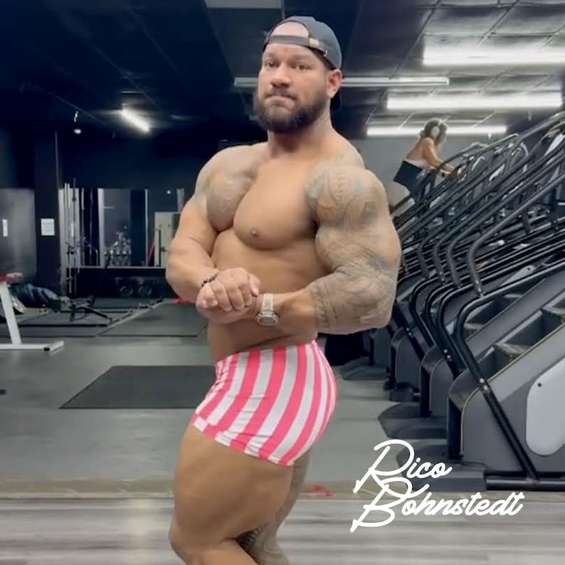 May include: A muscular person wearing pink and white striped shorts and a dark baseball cap poses in a gym. The person has tattoos on their arms and a watch on their wrist. The gym has exercise equipment and a person on a stair stepper.