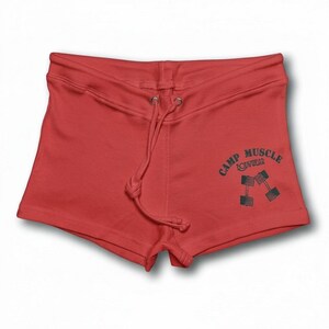 Puede incluir: Pantalones cortos deportivos rojos con cintura de cord&oacute;n y un gr&aacute;fico verde oscuro en la pierna derecha. El gr&aacute;fico muestra el texto CAMP MUSCLE BODYWEAR dispuesto en arco sobre una ilustraci&oacute;n de dos mancuernas cruzadas.