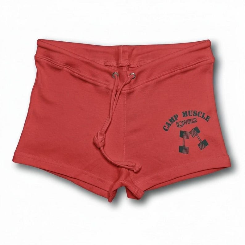 Puede incluir: Pantalones cortos deportivos rojos con cintura de cord&oacute;n y un gr&aacute;fico verde oscuro en la pierna derecha. El gr&aacute;fico muestra el texto CAMP MUSCLE BODYWEAR dispuesto en arco sobre una ilustraci&oacute;n de dos mancuernas cruzadas.