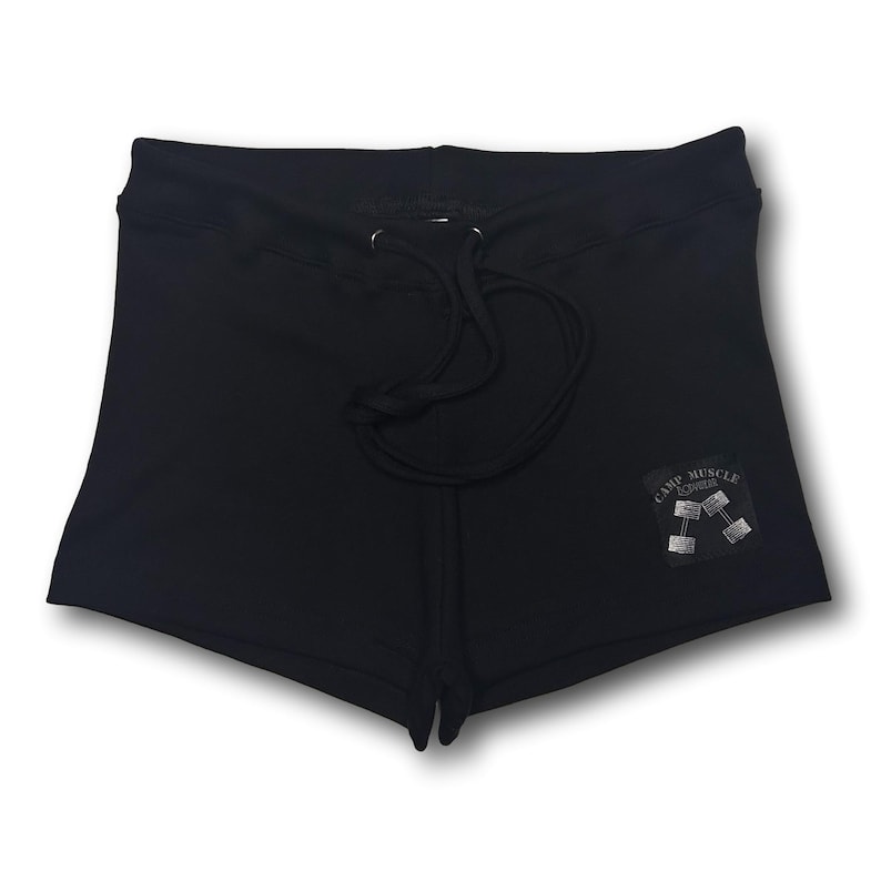 Puede incluir: Pantalones cortos deportivos negros con cintura de cord&oacute;n y un parche cuadrado en la pierna derecha. El parche muestra el texto "CAMP MUSCLE" sobre una ilustraci&oacute;n de dos mancuernas. Los pantalones tienen un dise&ntilde;o negro liso y sencillo.