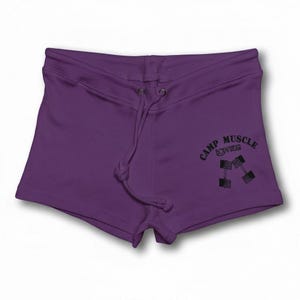 Puede incluir: Shorts de color morado oscuro con cintura de cord&oacute;n y ojales met&aacute;licos. La pierna derecha muestra un estampado gr&aacute;fico negro de dos mancuernas y el texto CAMP MUSCLE BODYWEAR en una fuente curva.