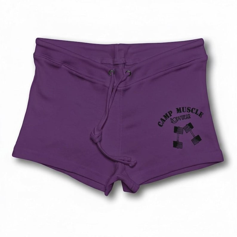Puede incluir: Shorts de color morado oscuro con cintura de cord&oacute;n y ojales met&aacute;licos. La pierna derecha muestra un estampado gr&aacute;fico negro de dos mancuernas y el texto CAMP MUSCLE BODYWEAR en una fuente curva.