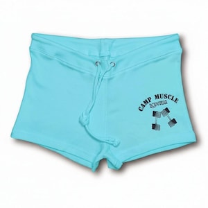 Puede incluir: Pantalones cortos deportivos de color azul claro con cintura de cord&oacute;n y ojales plateados. En la parte delantera derecha presenta un estampado gr&aacute;fico negro de dos mancuernas y el texto "CAMP MUSCLE BODYWEAR" en una fuente sans-serif.