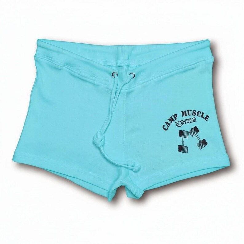Puede incluir: Pantalones cortos deportivos de color azul claro con cintura de cord&oacute;n y ojales plateados. En la parte delantera derecha presenta un estampado gr&aacute;fico negro de dos mancuernas y el texto "CAMP MUSCLE BODYWEAR" en una fuente sans-serif.
