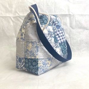 May include: A patchwork tote bag with a blue and white floral pattern and a navy blue strap. The bag has a small gingham check pocket on the front.