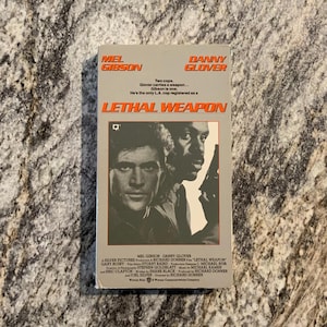 May include: A rectangular VHS tape case for the film Lethal Weapon, featuring a grey cover with orange text, the names Mel Gibson and Danny Glover, and a black and white portrait of the two actors, one holding a handgun, with production credits.