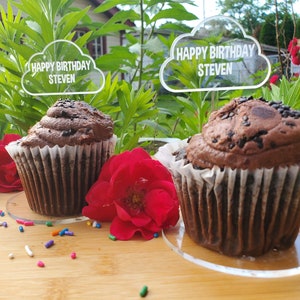 May include: Three chocolate cupcakes with chocolate chips on top, each topped with a clear acrylic cloud-shaped cake topper that says "Happy Birthday Steven". The cupcakes are on white cake stands and are surrounded by colorful sprinkles on a wooden surface.