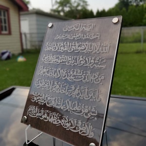 May include: A brown wooden plaque with a silver inscription of Arabic text. The text reads "Ayah Al-Kursi" and includes the first verse of the Quran. The plaque is mounted on a clear acrylic stand.