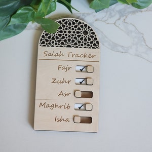 Puede incluir: Un rastreador de Salah de madera con un diseño de arco decorativo. El rastreador tiene cinco secciones para marcar las oraciones: Fajr, Zuhr, Asr, Maghrib e Isha. Cada sección tiene una casilla de verificación y un control deslizante.