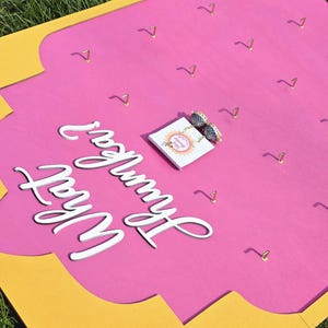 May include: A pink and yellow jewelry display board with gold-colored hooks. The board has the words "What Hummer Wear" in white script. A small card with earrings is placed on the board.