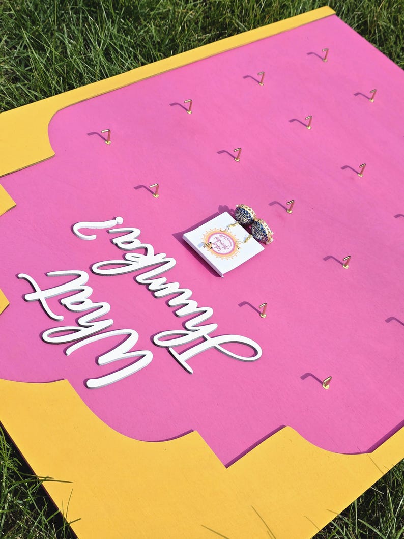 May include: A pink and yellow jewelry display board with gold-colored hooks. The board has the words "What Hummer Wear" in white script. A small card with earrings is placed on the board.