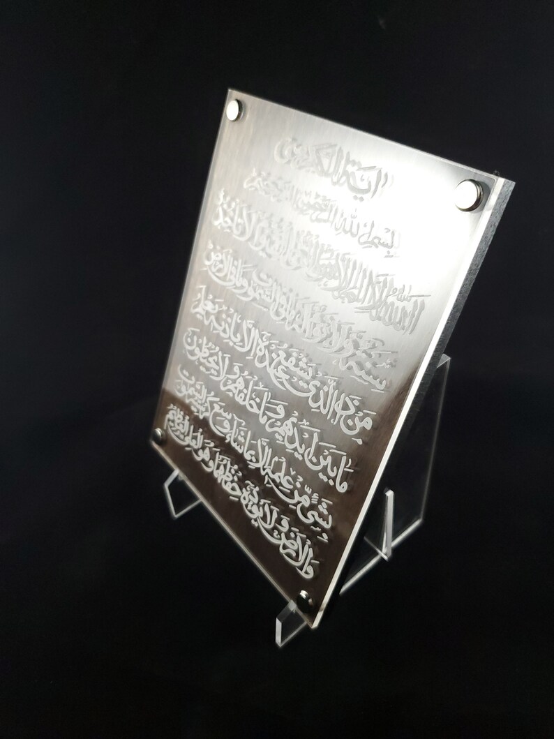 May include: A silver acrylic plaque with an engraved Islamic prayer in Arabic script. The plaque is displayed on a clear acrylic stand.