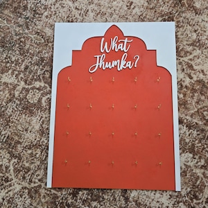 May include: A decorative wall hanging with a white border and a red center. The red center features small gold hooks and the white text "What Jhumka?" in a script font. The design is reminiscent of an arch.