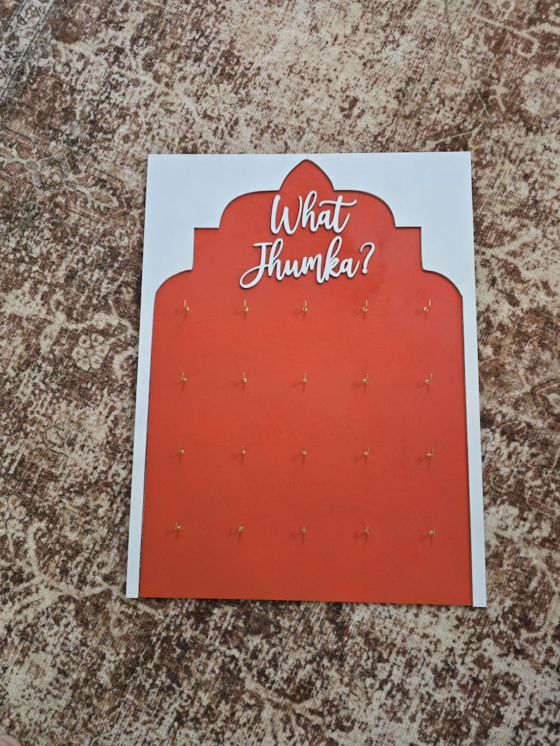 May include: A decorative wall hanging with a white border and a red center. The red center features small gold hooks and the white text "What Jhumka?" in a script font. The design is reminiscent of an arch.