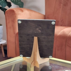 May include: A dark brown wooden picture frame with a light brown wooden easel stand. The frame is standing on a glass table.