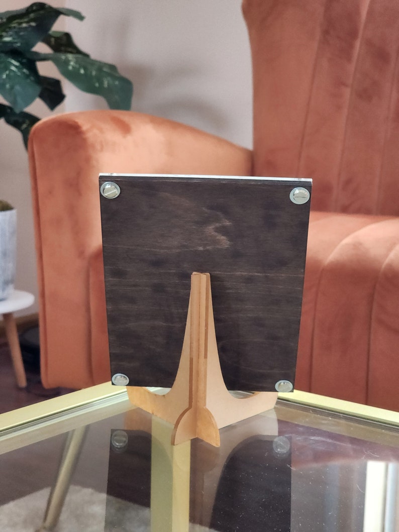May include: A dark brown wooden picture frame with a light brown wooden easel stand. The frame is standing on a glass table.
