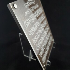 May include: A clear acrylic plaque with a brown wood-like background. The plaque features white Arabic calligraphy. The plaque is standing on a clear acrylic stand.