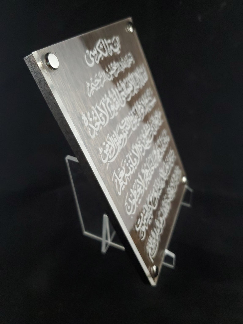 May include: A clear acrylic plaque with a brown wood-like background. The plaque features white Arabic calligraphy. The plaque is standing on a clear acrylic stand.