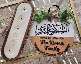 May include: A wooden round sign with a white top half and a brown bottom half. The top half features Arabic calligraphy that reads "Assalamu alaykum", which translates to "Peace be upon you". The bottom half features the text "Peace be upon you The Usman Family Welcomes You" in black lettering. The sign is decorated with a green leafy garland and a brown bow.