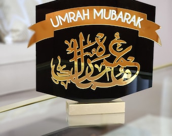 Peut inclure: Une plaque en acrylique noire avec des lettres dorées qui disent "Umrah Mubarak" et de la calligraphie arabe. La plaque est sur un support doré.