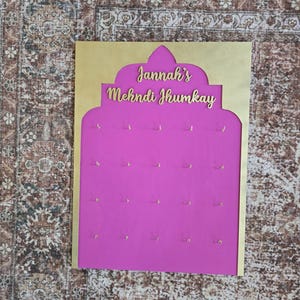 May include: A display board with a gold-colored frame and a bright pink interior. The text "Jannah's Mehndi Jhumkay" is displayed in gold lettering. Small gold hooks are evenly spaced across the pink surface, likely for hanging jewelry.