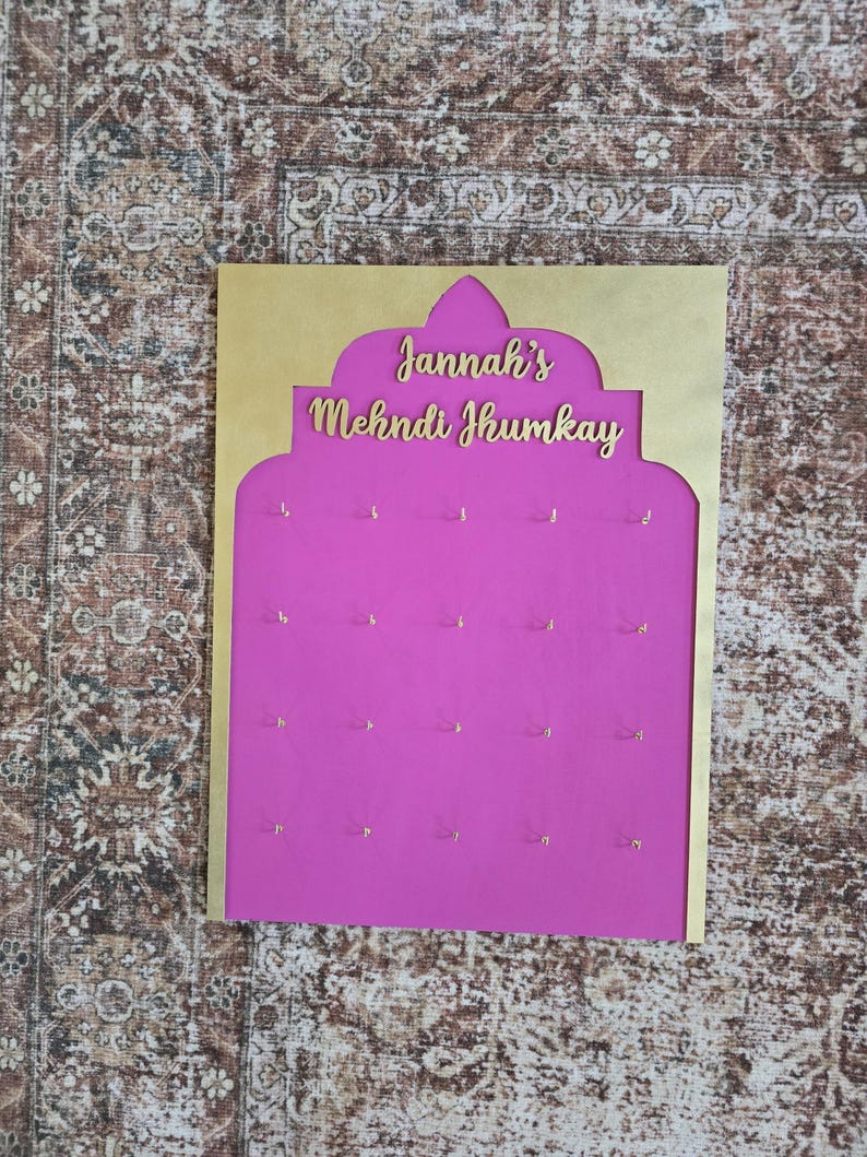 May include: A display board with a gold-colored frame and a bright pink interior. The text "Jannah's Mehndi Jhumkay" is displayed in gold lettering. Small gold hooks are evenly spaced across the pink surface, likely for hanging jewelry.