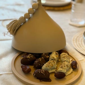 May include: A beige ceramic dome with a tassel and beads on top, open to reveal a plate of chocolate-covered dates and dates with white chocolate and nuts.