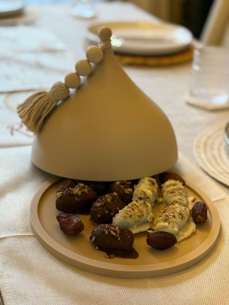 May include: A beige ceramic dome with a tassel and beads on top, open to reveal a plate of chocolate-covered dates and dates with white chocolate and nuts.