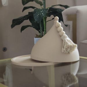 May include: A beige ceramic lidded dish with a wooden knob and tassel. The dish is on a glass table top.