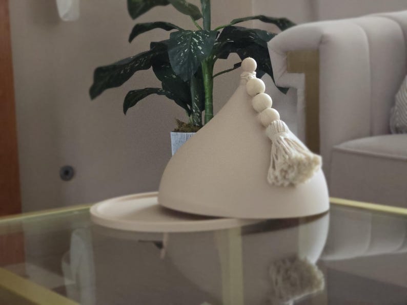 May include: A beige ceramic lidded dish with a wooden knob and tassel. The dish is on a glass table top.