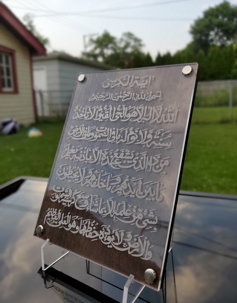 May include: A brown wooden plaque with a silver inscription of Arabic text. The text reads "Ayah Al-Kursi" and includes the first verse of the Quran. The plaque is mounted on a clear acrylic stand.