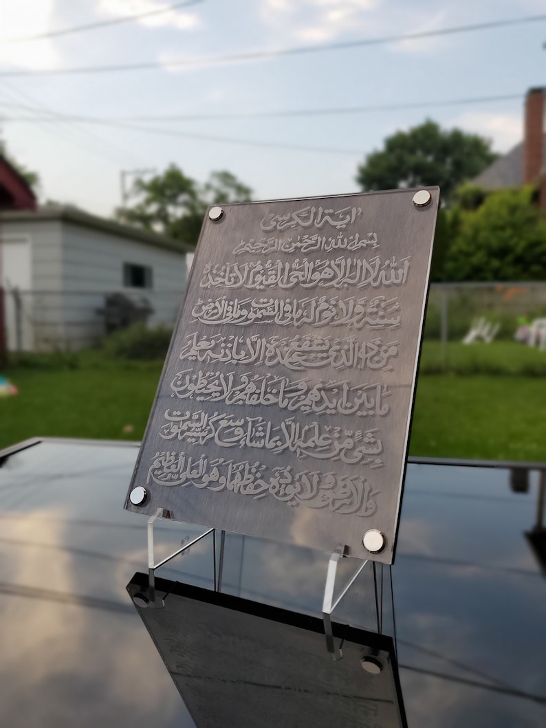 May include: A dark gray acrylic plaque with Arabic calligraphy in silver. The plaque is on a clear acrylic stand. The text reads "Ayah Al-Kursi" and includes the first verse of the Quran, "Bismillah ar-Rahman ar-Rahim".