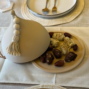 May include: A beige ceramic dome with a tassel and wooden beads, open to reveal a plate of dates and white chocolate covered dates. The plate has a beige background and the text "Ramadan Kareem" is written on a white runner.