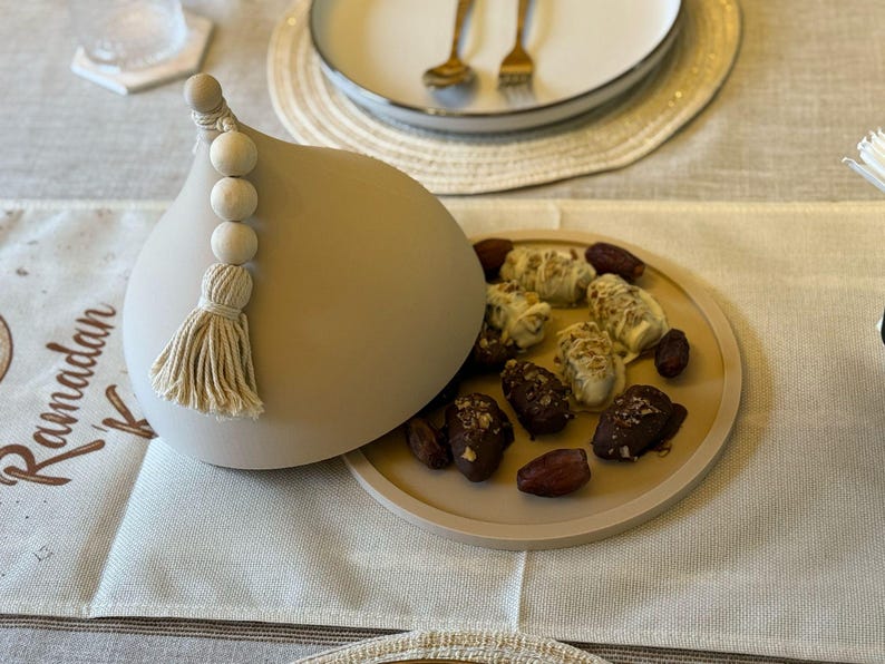 May include: A beige ceramic dome with a tassel and wooden beads, open to reveal a plate of dates and white chocolate covered dates. The plate has a beige background and the text "Ramadan Kareem" is written on a white runner.