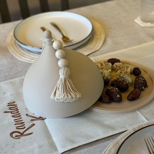 May include: A light gray ceramic dome with a white tassel and wooden beads. The dome is sitting on a table with a white tablecloth and a plate of dates and other treats.