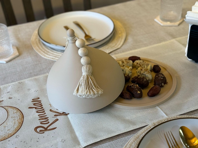 May include: A light gray ceramic dome with a white tassel and wooden beads. The dome is sitting on a table with a white tablecloth and a plate of dates and other treats.