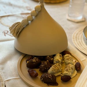 May include: A beige ceramic dome with a tassel and white beads on top, open to reveal a plate of dates covered in chocolate and white chocolate.