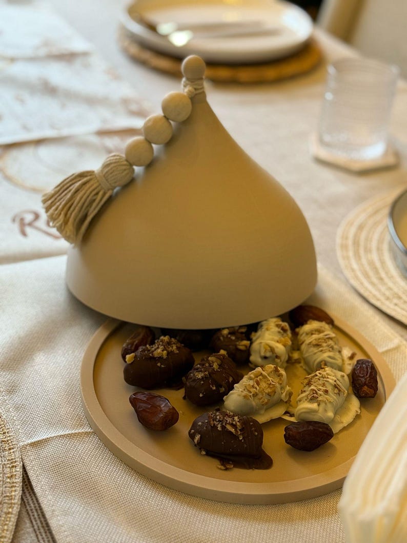 May include: A beige ceramic dome with a tassel and white beads on top, open to reveal a plate of dates covered in chocolate and white chocolate.