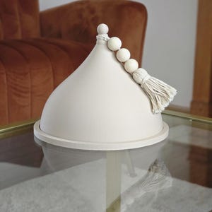 May include: A white ceramic dome with a wooden bead and tassel accent. The dome is sitting on a glass table.