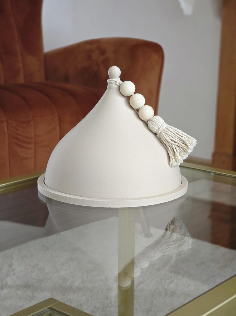 May include: A white ceramic dome with a wooden bead and tassel accent. The dome is sitting on a glass table.