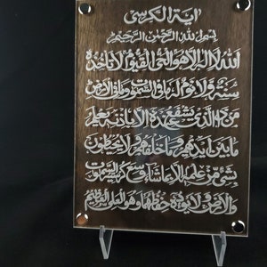 May include: A brown wooden plaque with white Arabic calligraphy. The text reads "Ayah Al-Kursi" and includes the first verse of the Surah Al-Baqarah.