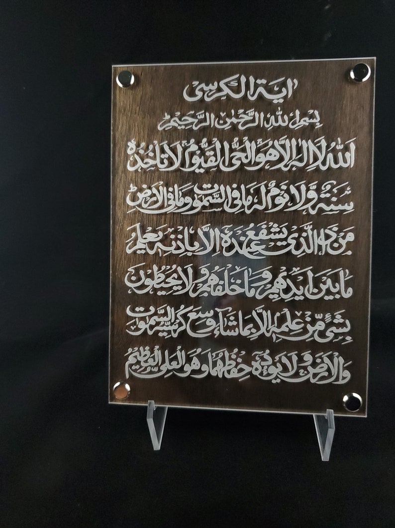 May include: A brown wooden plaque with white Arabic calligraphy. The text reads "Ayah Al-Kursi" and includes the first verse of the Surah Al-Baqarah.